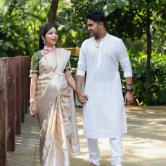 An expecting couple walks hand-in-hand during their outdoor baby shower photoshoot. They are dressed in elegant traditional Indian attire, with the mom-to-be in a beautiful off-white saree.