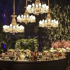 A glamorous bar setup for a cocktail night or reception. The curved black bar is placed under a canopy of hanging flowers and multiple crystal chandeliers, creating a luxurious and enchanting focal point.