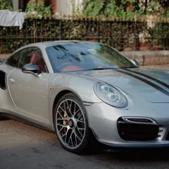 A front-facing view of a silver Porsche 911, highlighting its iconic headlights and aerodynamic design. The reflection of trees in the windshield adds a touch of the urban environment.