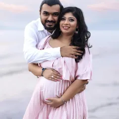 A classic couple pose during a serene beach photoshoot. He holds her from behind as they both smile at the camera, standing in the gentle waves with a beautiful sunset sky.