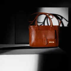 A classic tan handbag shot with dramatic, high-contrast lighting. The play of light and shadow accentuates the bag's structure and details.