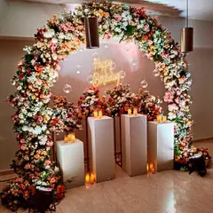 A slightly different angle of the full floral arch, showing the depth and texture of the flower arrangement. The setup is complemented by candles on plinths and a central neon sign.