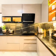 A showcase of modern luxury, this L-shaped kitchen features high-gloss beige cabinets paired with a dramatic black marble countertop and backsplash. We integrated under-cabinet lighting and glass-front display units to add depth and highlight the client's crockery.