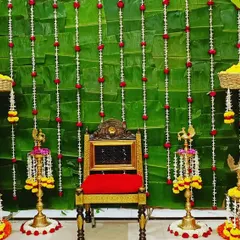 A closer look at a traditional decor setup for a home ceremony. The backdrop of fresh green leaves is adorned with strings of white and red flowers, complemented by brass lamps and baskets of marigolds.