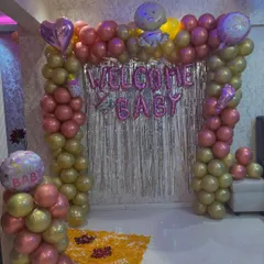 A joyous 'Welcome Baby' decoration for a new arrival at home. The setup features a balloon arch in pink and gold, a silver fringe backdrop, and 'Welcome Baby' foil balloons.
