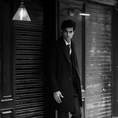 A moody, low-light shot of the model in a suit and overcoat, leaning against a dark doorway. The single light source creates a dramatic, high-contrast look.