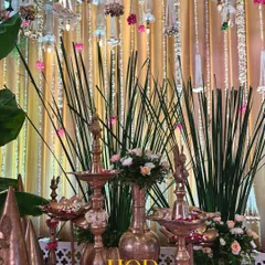 A collection of traditional brass lamps (diyas) and vessels, interspersed with fresh flowers and greenery, creating a rich and authentic decor corner.