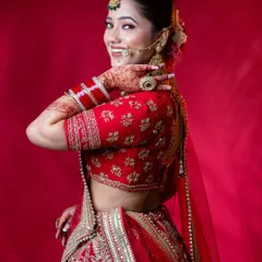 A vibrant and joyful bridal shot against a solid red background. The model's happy expression and pose showcase the complete look, including the embroidered red lehenga, traditional jewellery, and intricate henna design, perfect for demonstrating a full bridal styling.