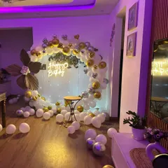 A sophisticated birthday ring decoration in a home setting. The white, gold, and silver balloon garland is paired with a neon sign and fairy lights for a warm, celebratory glow.