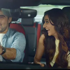 A fun, energetic shot from inside the car. The woman's bright yellow top and the man's casual blue shirt create a vibrant, happy mood for their road trip.