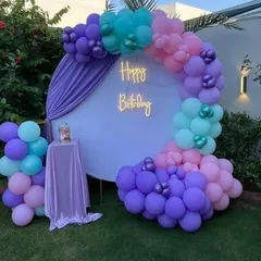 An outdoor birthday decoration featuring a circular backdrop with a neon "Happy Birthday" sign, framed by a beautiful balloon garland in purple, pink, and teal.
