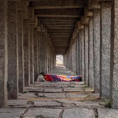 A person sleeps in the long, pillared corridor of a temple, sharing the space with squirrels. This candid shot captures a quiet moment of daily life persisting within the historic ruins.