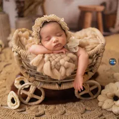 A close-up of the baby in the rustic wicker pram, showing the classic newborn pose with hands tucked under the chin. The soft, chunky knit adds another layer of texture.