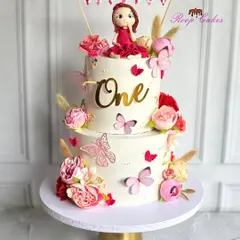 A beautiful two-tier cake celebrating "One." This whimsical design is decorated with pink and red flowers, butterflies, and a handcrafted doll topper, making it a perfect centerpiece for a first birthday.