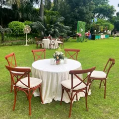 Simple and elegant outdoor seating for a daytime event. The wooden cross-back chairs and clean white tablecloths create a rustic and sophisticated look on a lush green lawn.