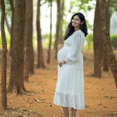 A full-length solo portrait in a peaceful woodland setting. The mother-to-be stands confidently, cradling her bump in a lovely white dress, showcasing the simple elegance of pregnancy.