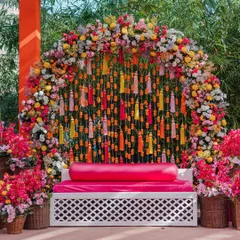 A vibrant and colorful Mehendi seating area, featuring a backdrop of hanging tassels and marigolds, framed by a lush arch of multi-colored flowers. This decor is full of joy and perfect for a festive celebration.