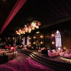 A dramatic lounge area with a dark, moody atmosphere. The space features plush pink sofas on a tiered platform, gothic-style arched windows, and a ceiling adorned with hanging flowers and lanterns.