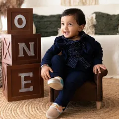 A one-year-old boy dressed in a smart suit and hat smiles while sitting next to "ONE" blocks, capturing a classic first birthday portrait.