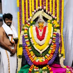 I stand next to my work, a fully decorated deity at the Vasavi Matha Temple, adorned with dense, colorful flower garlands.