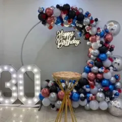 A stylish and modern setup for a 30th birthday. This includes a circular frame with a balloon arch in black, silver, red, and blue, a 'Happy Birthday' neon sign, and large light-up marquee numbers for the age.