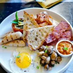 The full English breakfast, CAARA style. A plate loaded with a sunny-side-up organic egg, bacon, chicken sausages, sautéed mushrooms, baked beans, a grilled tomato, and artisanal sourdough toast.