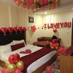 A full view of a classic romantic room setup, featuring red and gold ceiling balloons, "I LOVE YOU" foil letters, and rose petals on the bed and floor.
