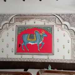 A classic Pichwai style painting, this mural brings serenity and tradition into the home. Featuring a beautifully adorned cow against a vibrant red backdrop, framed by intricate Rajasthani architectural motifs, it is a piece of heritage art that creates a divine atmosphere.