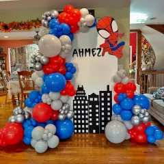 A Spider-Man themed birthday party setup. This decoration includes a city skyline backdrop, a Spider-Man cutout, and a balloon arch in the classic red, blue, and silver colors.