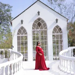 A beautiful solo portrait of a mother-to-be in a red gown, standing on a white bridge in front of a charming chapel-like building.