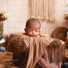 This newborn is posed sleeping on top of a textured log prop. The background features a white brick wall, macrame art, and dried pampas grass for a complete boho look.