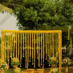 This minimalist Haldi setup features a clean backdrop of marigold strings, creating a vibrant wall of yellow. The traditional brass urlis are ready for the ceremony, blending simplicity with cultural richness.