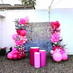 A vibrant birthday setup featuring a blue and silver shimmer wall. The backdrop is framed with bright pink and silver balloons, including starburst foil balloons, and includes pedestals for cake or props.
