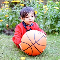 A young boy in a red suit jacket holds a basketball during a sports-themed outdoor photoshoot in a garden.