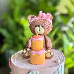 A detailed shot of the adorable fondant teddy bear topper, complete with a pink bow and a tiny two-tier cake with a candle.