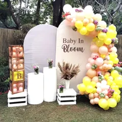 "Baby in Bloom" is such a beautiful theme for a baby shower. This outdoor setup features a lovely balloon arch in pastel yellows and pinks, elegant white pedestals, and custom "BABY" blocks, all set against a lush garden backdrop.