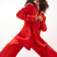 A dynamic, wide-stance pose in the same red power suit. This image captures a sense of movement and attitude, highlighting the fluid tailoring of the oversized blazer and trousers for a bold editorial statement.