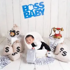 A series from a 'Boss Baby' themed newborn shoot. The setup includes a tiny armchair, a miniature briefcase, and prop money, creating a playful and humorous scene for a baby boy's first portraits.