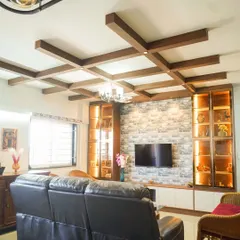 This living room showcases a coffered ceiling with wooden rafters and integrated display units. The design balances a stone-clad TV wall with warm, backlit shelving, creating a space that is both grand and inviting.