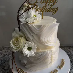 An elegant two-tier birthday cake featuring delicate white wafer paper ruffles and fresh white flowers. The design is complemented by rustic chocolate branches for a touch of nature.