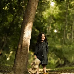 A smiling girl with curly hair holds a teddy bear while standing by a tree in a sun-dappled forest. The natural lighting and a butterfly in motion create a whimsical and heartwarming childhood portrait.