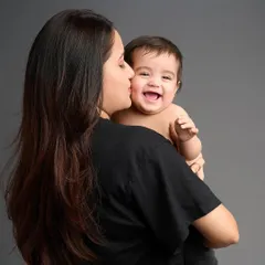 A candid moment of a mother kissing her laughing baby's cheek. The simple grey backdrop makes their joyful interaction the sole focus, resulting in a powerful and emotional portrait.