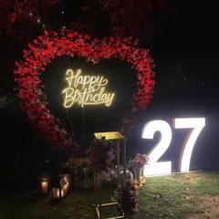 A romantic outdoor decoration featuring a large heart-shaped frame made of red flowers. This setup, shown here for a 27th birthday, is also perfect for a surprise proposal or anniversary celebration, complete with a custom neon sign and marquee numbers.