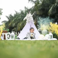 For outdoor pre-birthday shoots, we create beautiful picnic-style setups. This one features a decorated teepee, marquee letters spelling 'ONE', and atmospheric props like lanterns and colored smoke for a touch of magic.