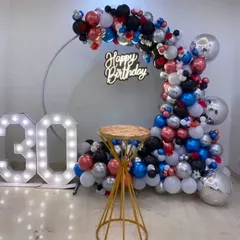 A vibrant 30th birthday party decoration. A circular frame is adorned with a colorful mix of red, blue, black, and silver balloons, complemented by a neon sign and light-up '30' numbers.