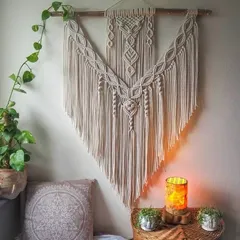 The same V-shaped design styled in a cozy corner with plants and a warm lamp. The intricate knotwork and long fringe create a peaceful and inviting atmosphere.