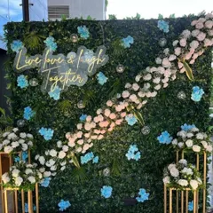 A modern and fresh selfie backdrop with a green foliage wall, accented with blue and white flowers and a "Live, love & laugh together" neon sign.