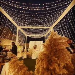 A cozy lounge area for a boho-themed event at night. A canopy of fairy lights illuminates the space, with large pampas grass arrangements adding texture and warmth.