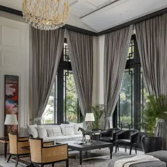 Double-height windows in the living room of the House of Verandahs flood the space with natural light and frame views of the surrounding greenery. We used a palette of muted tones and custom furniture to create an atmosphere of both grandeur and comfort.