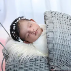 A close-up of a sleeping baby in a vintage-style grey wicker pram. The pearl headband adds a touch of classic elegance to this simple and sweet portrait.